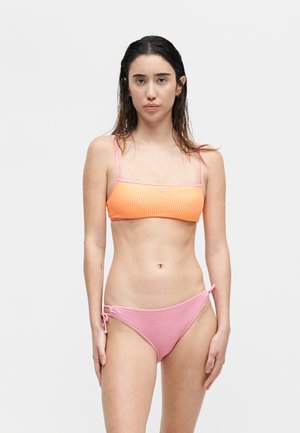 Young woman with wet hair wearing an orange bandeau bikini top and pink textured bikini bottoms, standing against a plain white background.