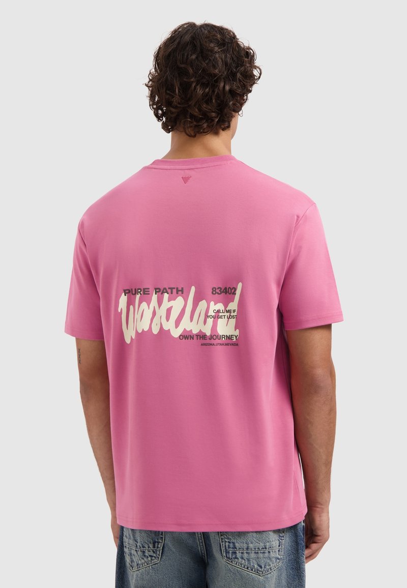 Mann mit lockigem, dunklem Haar, der ein pinkfarbenes T-Shirt mit "Westplay" und reisebezogenen Text trägt, kombiniert mit blauen Denim-Jeans, von hinten gezeigt.