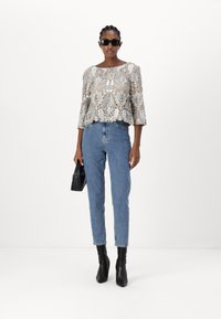 Spitzenoberteil mit floralem Muster, in hellbeigen und silbernen Tönen, mit Glockenärmeln; kombiniert mit hochgeschnittenen blauen Jeans und schwarzen Knöchelstiefeln.