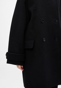 Cappotto doppiopetto nero in lana, con grandi bottoni, tasche laterali e polsini pieghettati. Texture liscia con una silhouette ben strutturata.