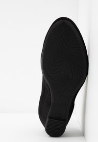 Chaussure en suede noir avec une semelle en caoutchouc texturé présentant un motif ondulé ; le talon est arrondi et le nom de la marque "Gabor" est visible.