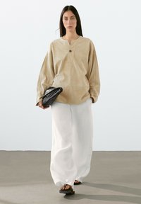 Beiger Wildlederpullover mit einem halben Knopfverschluss, weiten Ärmeln, kombiniert mit lockeren weißen Leinenhosen und schwarzen Flip-Flops, während sie eine strukturierte schwarze Clutch hält.