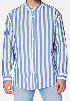 Mand iført langærmet skjorte med lodrette blå og hvide striber, kombineret med lyseblå jeans, beskåret ved nakke og lår.