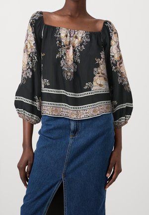 Femme portant un chemisier noir à fleurs, épaules dénudées avec manches bouffantes, et une jupe en denim bleu avec une fente à l'avant, se tenant devant un fond uni.