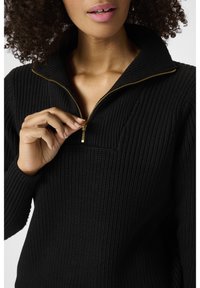 Schwarzer Pullover mit hohem Kragen und Reißverschluss, mit einem strukturierten Strickmuster und goldenem Reißverschluss-Akzent.