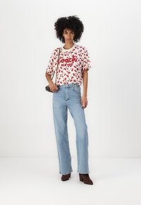 Tricou alb cu model de cireșe roșii și logo-ul "Coach", asortat cu blugi evazați de culoare albastră deschisă și cizme cu toc maro. Accesoriu: geantă tip crossbody.