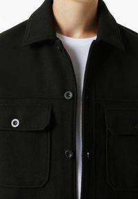 Černá vlna smíšená bunda s límcem, dvěma předními kapsami a matnými knoflíky. Tkanina má jemnou texturu. Pod ní bílé tričko.