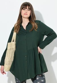 Camicia button-up verde scuro con design peplum, maniche lunghe e colletto, abbinata a una borsa intrecciata beige. Tessuto testurizzato e bottoni dorati.