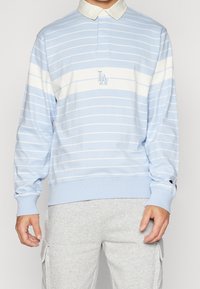 Světle modrá polokošile s dlouhým rukávem, bílými pruhy a bílým límcem, s vyšitým logem. Obsahuje žebrované manžety a volnější střih.