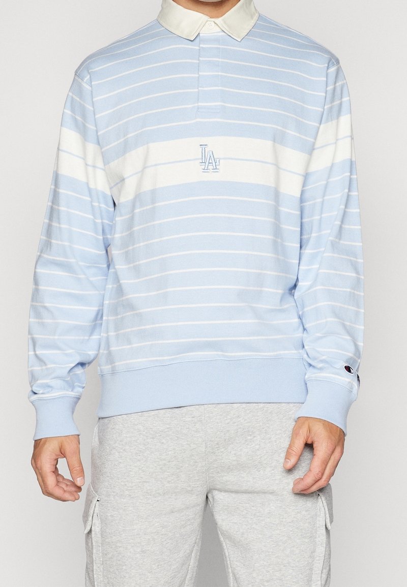 Světle modrá polokošile s dlouhým rukávem, bílými pruhy a bílým límcem, s vyšitým logem. Obsahuje žebrované manžety a volnější střih.