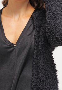 Black textured cardigan with a fuzzy material, featuring a v-neck top with a subtle clasp detail. The overall look is layered and soft.