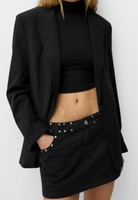 Svart skräddarsydd kavaj över en svart crop-top, tillsammans med en svart jeanskjol med metalliska nitar och en figurnära design.