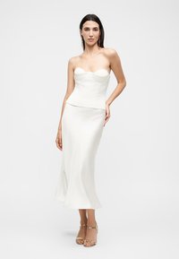 Femme portant un corset blanc sans bretelles et une longue jupe assortie, debout avec une main sur la hanche devant un fond uni.