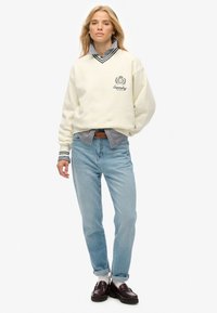 Sudadera color crema con detalles en verde, que presenta un logo. Camisa de botones a rayas por debajo, combinada con jeans de color azul claro y mocasines oscuros.