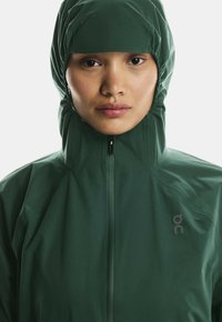 Roheline veekindel jakk kõrge kraega ja kapuutsiga, esisepal on lukku ja vasakul rinnal logo. Sile, kerge kangas.