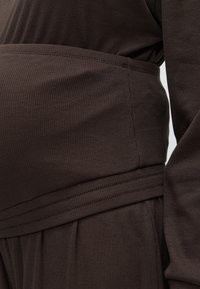 Κοντινή λήψη ενός σκουρόχρωμου καφέ ριμπ ύφανσης ενδύματος με ραμμένες ραφές, εστιασμένο στη μέση και την περιοχή του μανικιού.