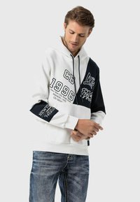 Kapuzensweatshirt mit einem Farbblockdesign in weißen und marineblauen Bereichen, textilem Grafikdruck und gerippten Bündchen. Kombiniert mit blauen Jeans.