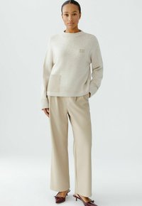 Beige Strickpullover mit lockerer Passform und einer Tasche, kombiniert mit weiten beigefarbenen Hosen und dunkelbraunen Mules, vor einem hellen Hintergrund.