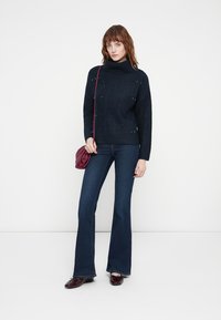 Pull en maille bleu foncé avec un col montant, présentant des motifs en diamant subtils et des accents. Assorti à un jean en denim évasé et un sac à main bordeaux.