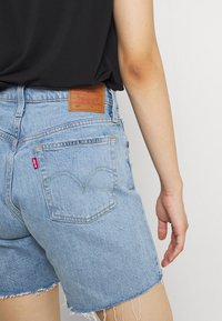 Pantalones cortos de mezclilla en azul claro, con dobladillos deshilachados, un diseño clásico de cinco bolsillos y una etiqueta de marca de cuero marrón en la parte trasera de la cintura.
