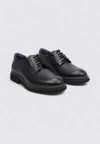 Une paire de chaussures de ville pour hommes en cuir noir, avec des lacets et des semelles robustes, présentées sur un fond gris clair uni.