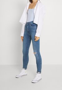 Un body azul claro debajo de una camiseta blanca corta, combinado con jeans ajustados azules que presentan detalles desgastados y zapatillas deportivas blancas de caña alta.