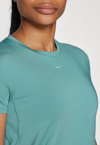 Teal atletisk t-shirt med rund halsringning och korta ärmar, med en liten ljusgrå Nike-logo på vänstra bröstet. Slät textur.