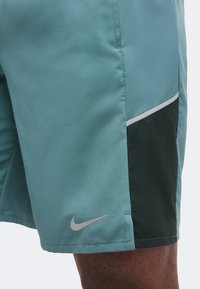 Τιρκουάζ αθλητικό σορτς με μαύρα διατρητά πλαϊνά πάνελ και ανακλαστικό λογότυπο Nike κοντά στο τελείωμα στον μηρό ενός ατόμου.