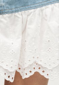 Λευκό ύφασμα με κεντημένα λουλουδάτα και γεωμετρικά σχέδια και κυματιστά άκρα κάτω από μπλε τζιν ύφασμα.