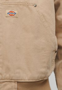 Τσέπη μπεζ καμβά σε μπουφάν με εμφανές έμβλημα της μάρκας Dickies και μεταλλική τρουκ στο πλαϊνό ράμμα.