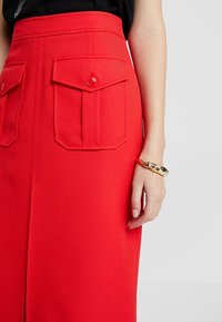Falda roja de tela texturizada con cintura alta, que presenta dos bolsillos frontales rectangulares y detalles de botones. Pulsera dorada en la muñeca.