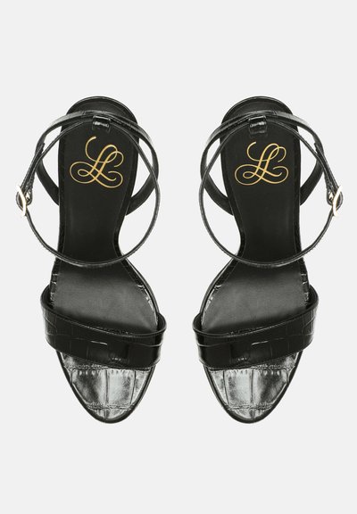 Sandalias de tacón negro con una textura brillante de grabado de cocodrilo, tiras delgadas y una tira en el tobillo. Logo dorado en la plantilla. Diseño de punta abierta.