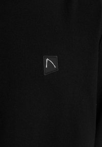 Schwarzes gestricktes Kleidungsstück mit einem kleinen, quadratischen schwarzen Aufnäher mit einem weißen abstrakten Design. Glatte Textur, keine sichtbaren Muster oder Verzierungen.