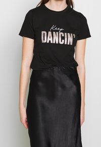 Camiseta de algodón negra con texto brillante que dice "Sigue bailando". Combinada con una falda negra de satén, de textura suave y diseño sencillo.