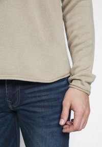 Beżowy sweter wykonany na drutach z długimi rękawami, o prążkowanej fakturze i zaokrąglonym brzegu, zestawiony z ciemnoniebieskimi dżinsami.