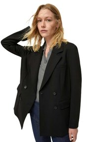 Blazer nero doppiopetto con vestibilità sartoriale, ampi revers e tasche laterali. Il modello indossa una camicia grigia sotto, abbinata a jeans casual.