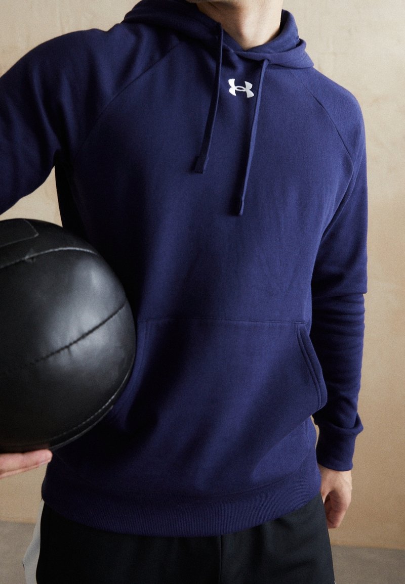 Personne portant un sweat à capuche bleu marine Under Armour, tenant un ballon médicinal en cuir noir sur un fond beige uni.
