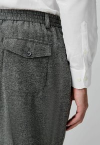Gray textured trousers featuring a back pocket with a black button and a waistband. Paired with a white button-up shirt.