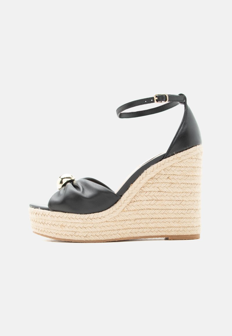 Μαύρο δερμάτινο σανδάλι πλατφόρμας με λουράκι στον αστράγαλο, δεμένο μπροστινό σχέδιο, χρυσή λεπτομέρεια και χοντρή πλεκτή σόλα πλατφόρμας.