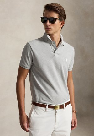Homme portant un polo gris clair à col relevé, pantalon blanc, ceinture marron, lunettes de soleil et une montre-bracelet sur un fond neutre.