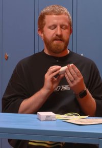 Wit cilindrisch product in handen gehouden, met een glad oppervlak. Een kleine rechthoekige doos staat op een blauwe tafel bedekt met papieren.