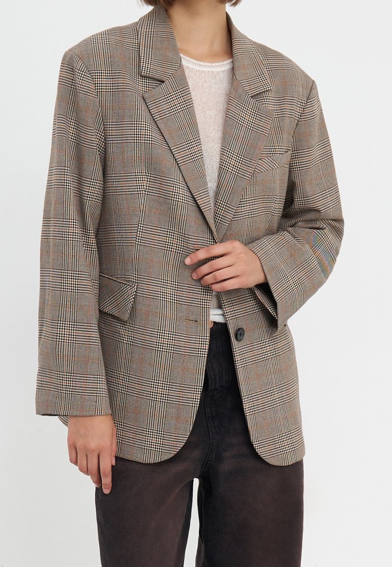 Personne portant un blazer oversized à carreaux beige et marron sur un top en maille blanc et un pantalon sombre, une main ajustant le blazer.