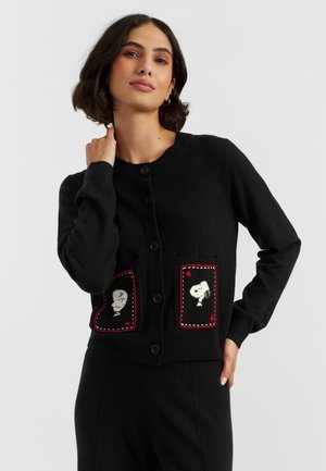 Eine Frau posiert in einer schwarzen Strickjacke mit Snoopy-Motiven auf den Taschen und zeigt einen stilvollen, verspielten Look vor einem schlichten Hintergrund.