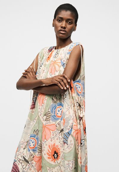Γυναίκα με κοντά μαλλιά που φοράει αμάνικο φόρεμα με λουλουδένιο μοτίβο σε παστέλ πορτοκαλί, μπλε και πράσινο, με σταυρωμένα τα χέρια και ουδέτερη έκφραση, σε απλό φόντο.
