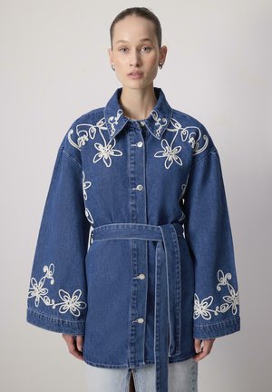 Nő kék farmer öves dzsekiben, fehér virágos hímzéssel a galléron, vállakon és bő ujjakon, egy egyszínű háttér előtt áll.