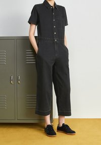 Svart denimjumpers med korta ärmar, framknäppning, bröstficka, vida ben och en slät yta. Matchad med svarta sneakers.