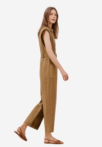 Brun linneoverall med korta ärmar, ett bälte i midjan och sidofickor, tillsammans med bruna sandaletter med remmar. Stående pose.