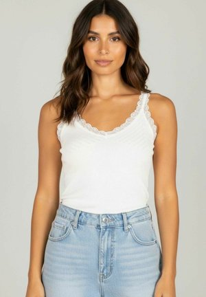 Femme aux cheveux bruns ondulés portant un débardeur blanc orné de dentelle et un jean taille haute bleu clair sur un fond clair.