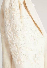 Abrigo de lana beige con intrincados bordados florales y detalles dorados. La textura es suave, e incluye un cuello con muesca y detalles de botones.