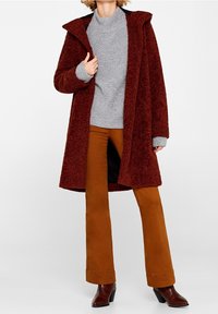 Manteau en fourrure bordeaux avec capuche, pull en maille gris et pantalon évasé beige. Caractéristiques : manches larges et coupe décontractée. Bottines marron complètent le look.